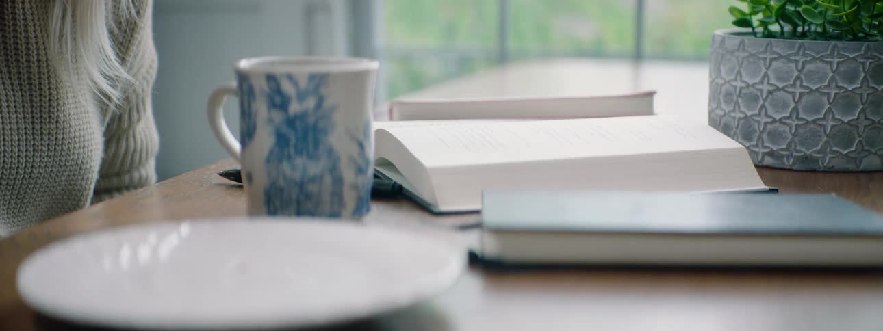 Female sits down at dining table in front of an open book and coffee and then picks up an iced cinnamon rolls from a dessert plate to take a bite and then places the uneaten portion back on the plate again