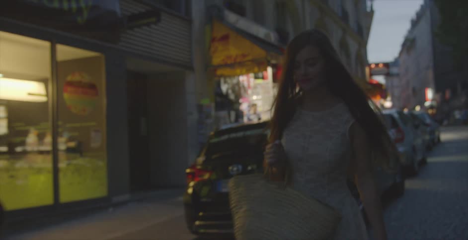 A woman carrying a large purse walks down a city street at dusk