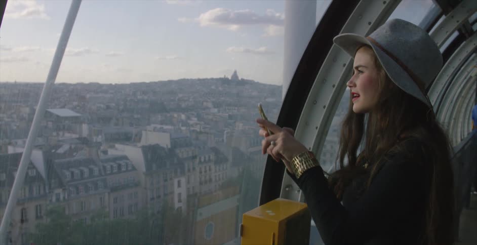 A woman stands by a window overlooking a city before taking some photos with her phone