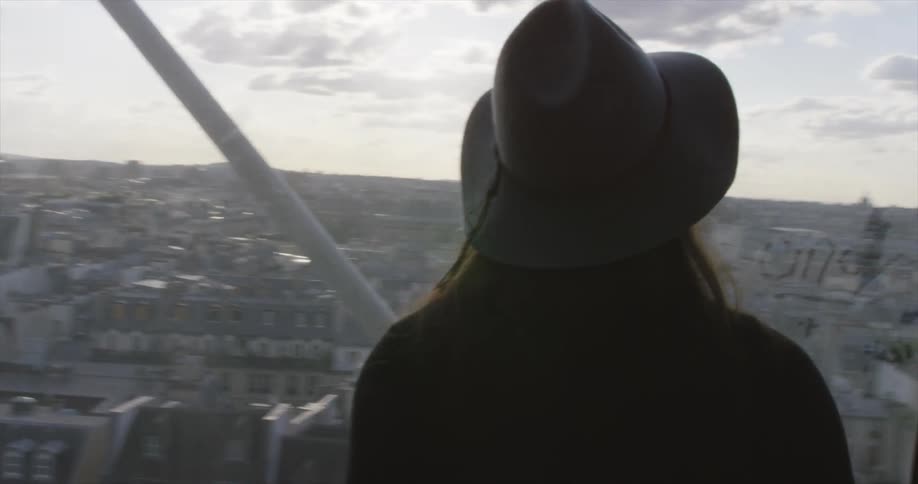 A woman walks over to a large window overlooking a city and smiles