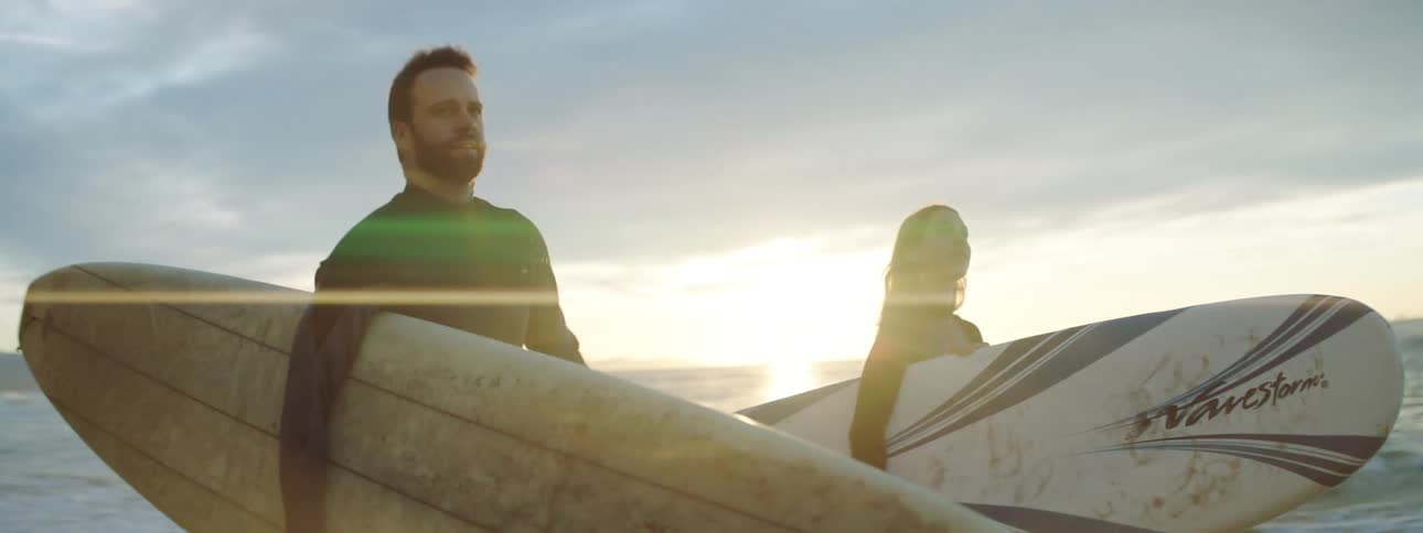 A man and woman walk along the shore carrying surfboards