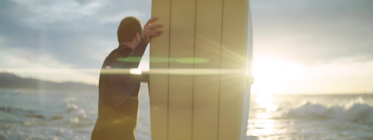 A man and woman laugh as they hold their surf boards and look out at the water