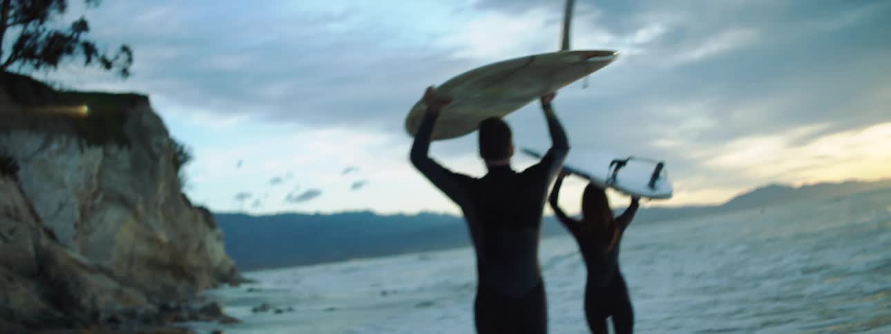 A man and woman carry surfboards above their hands as they walk along the beach beneath a low cliff