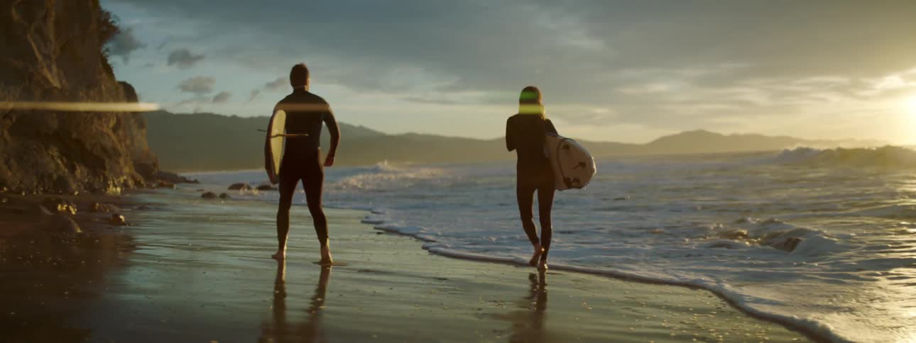 A man and woman walk down the beach carrying surf boards