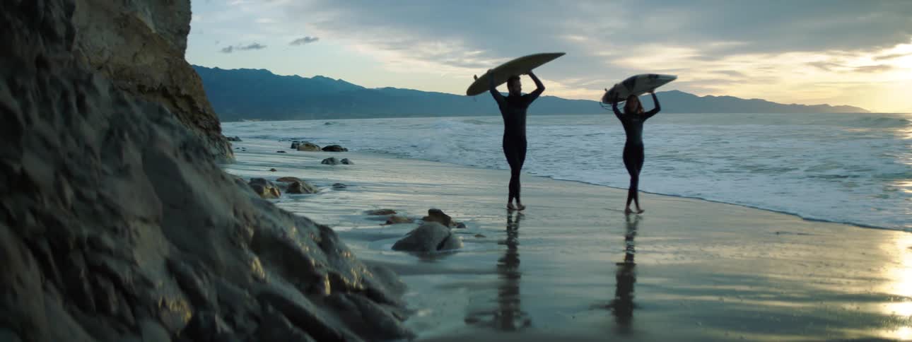 Two surfers balance surfboards on their heads on a sandy beach at sunset