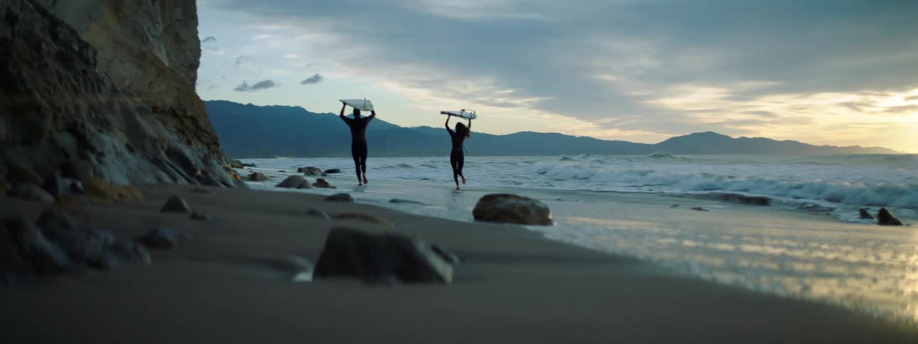 Man and woman each carry a surfboard on their heads as they walk along the beach