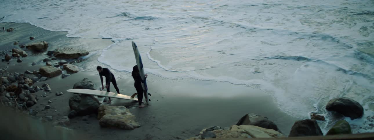 A man and woman in wet suits hold surfboards on a sandy beach