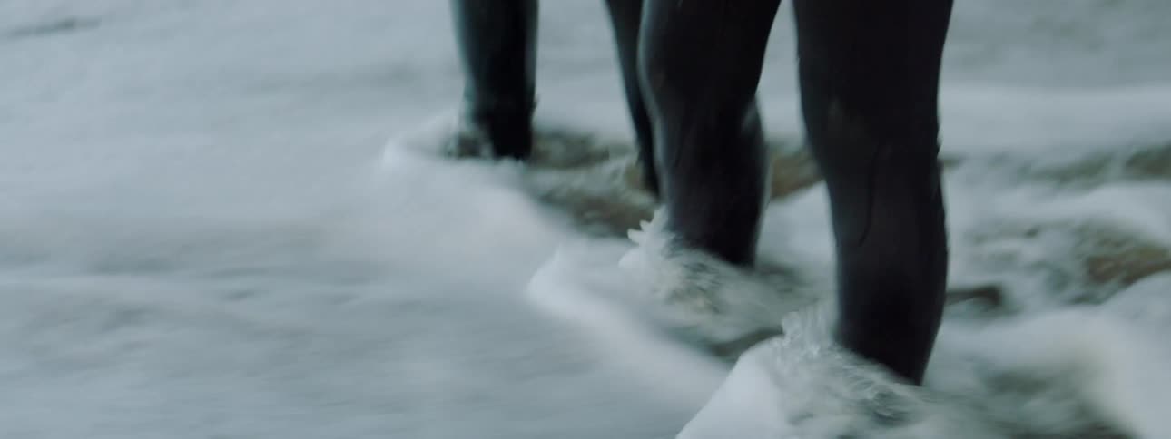 A man and woman in wet suits run in place on a wet beach