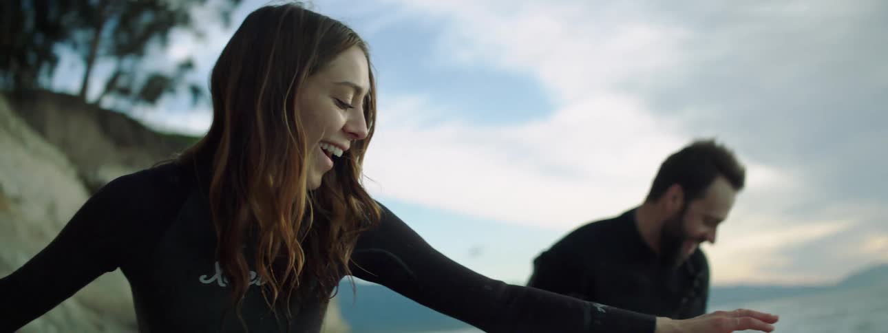 A woman in a wet suit practices surfing on a beach next to a man doing the same as a third man watches