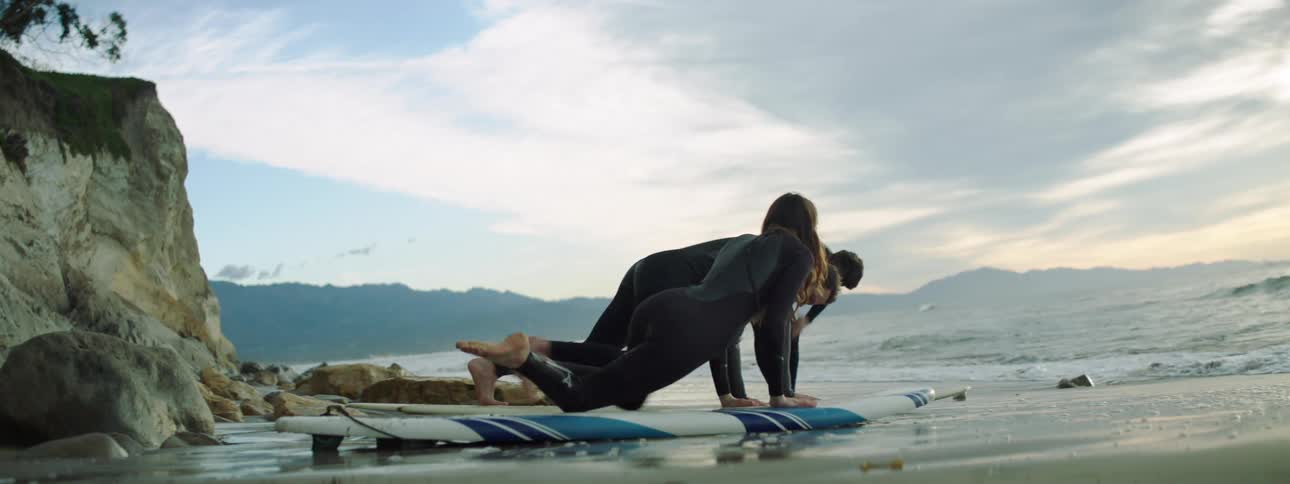 A man and woman lie down on surfboards on the beach as they listen to an instructor