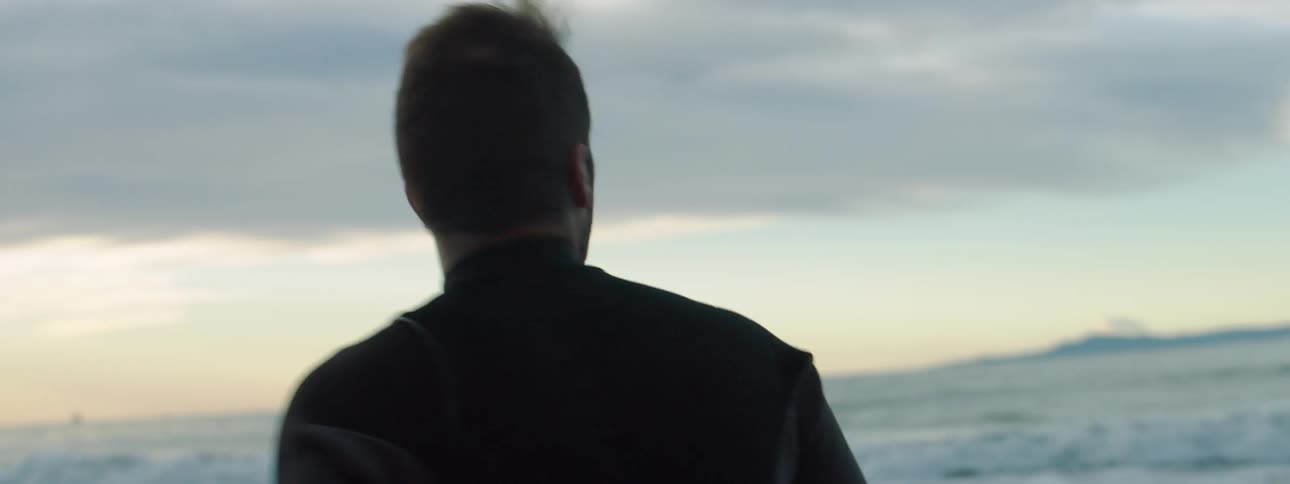 A man and woman wearing wet suits stand on a beach looking at the water