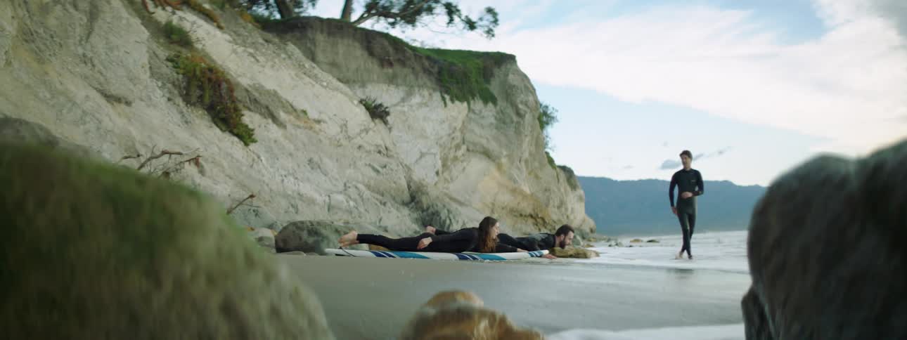 A man and woman lie on their surfboards on the beach practicing their paddling as an instructor watches