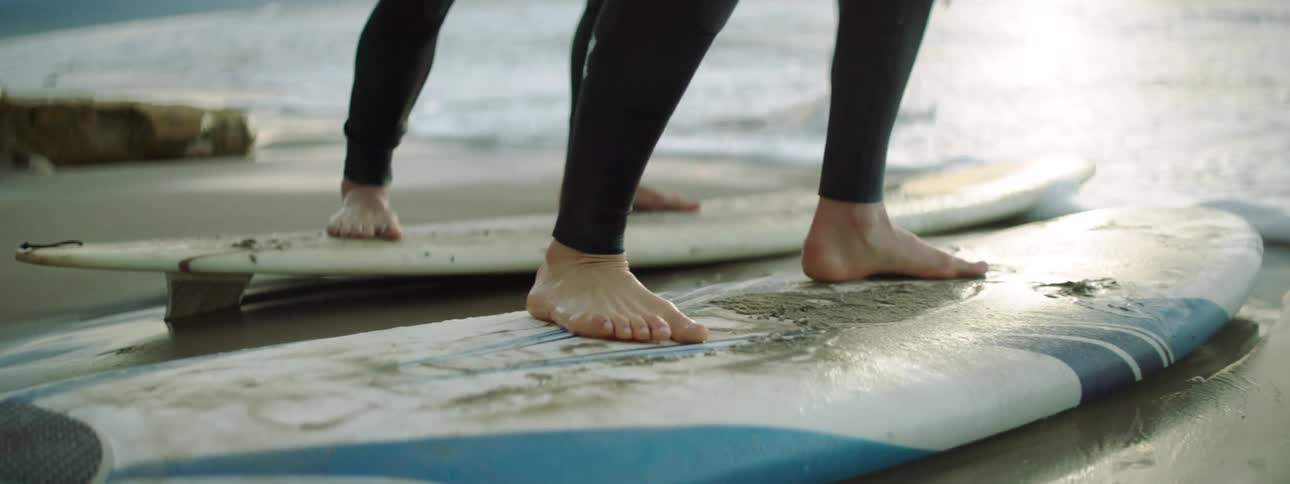 A male and female surfer practice rising from their bellies to a stand on their surfboards on a beach as the tide washes over their feet