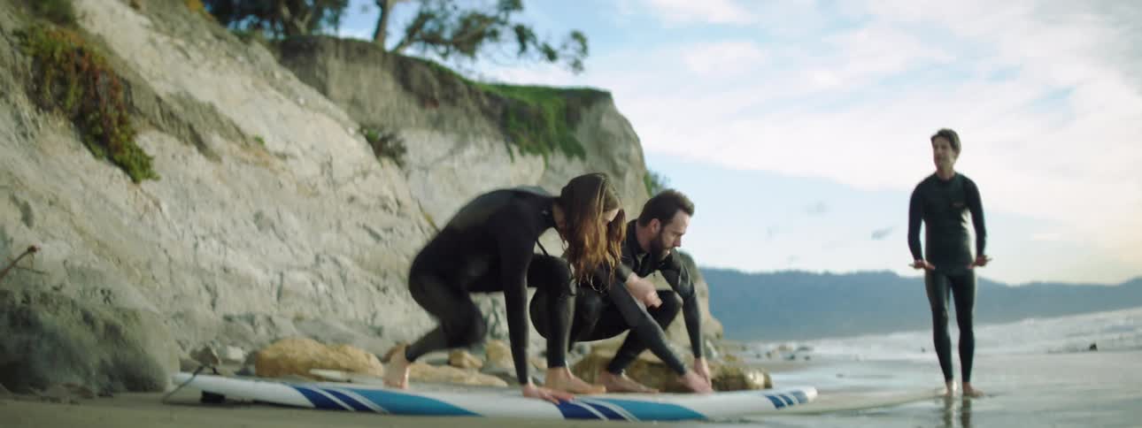 A man instructs a man and a woman on how to rise to a stand atop a surfboard