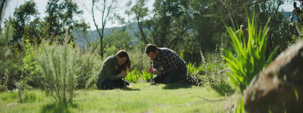 A man and woman try to make fire with sticks outdoors
