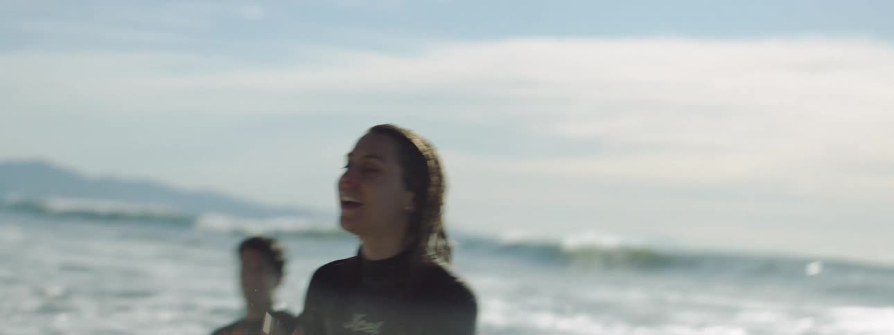 A man and woman stand in the ocean with surfboards as the woman calls out happily toward the coast