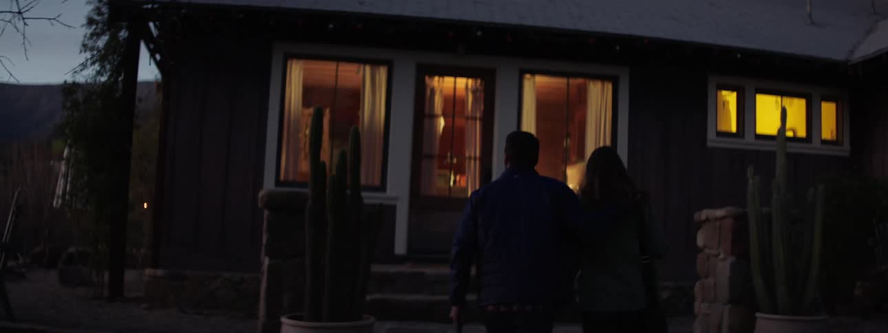 A man and a woman with bags walking into a house with trees and the sky in the background