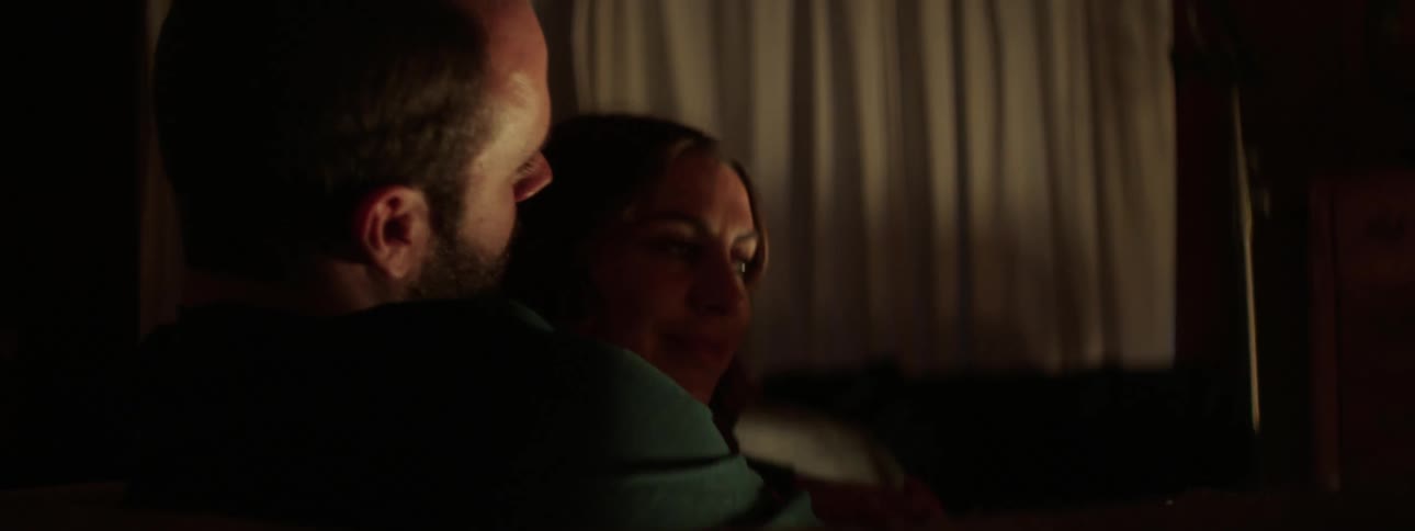 A man and a woman sitting on a couch hugging with a window and curtains in the background