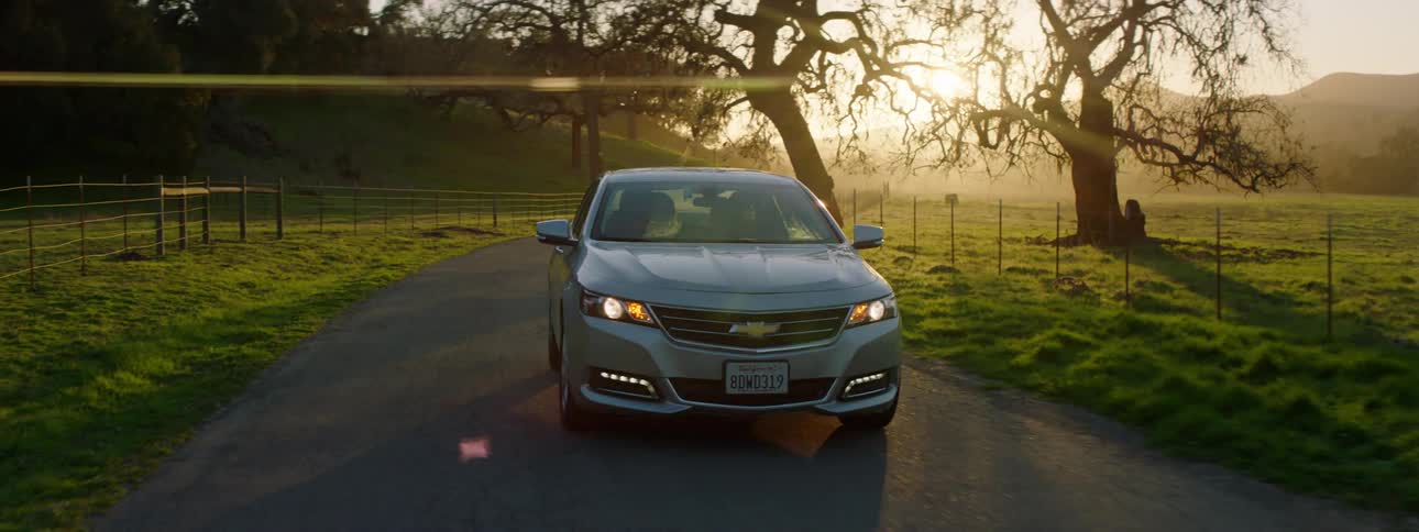 A car travels along a road between fences in the countryside as the sun shines from behind