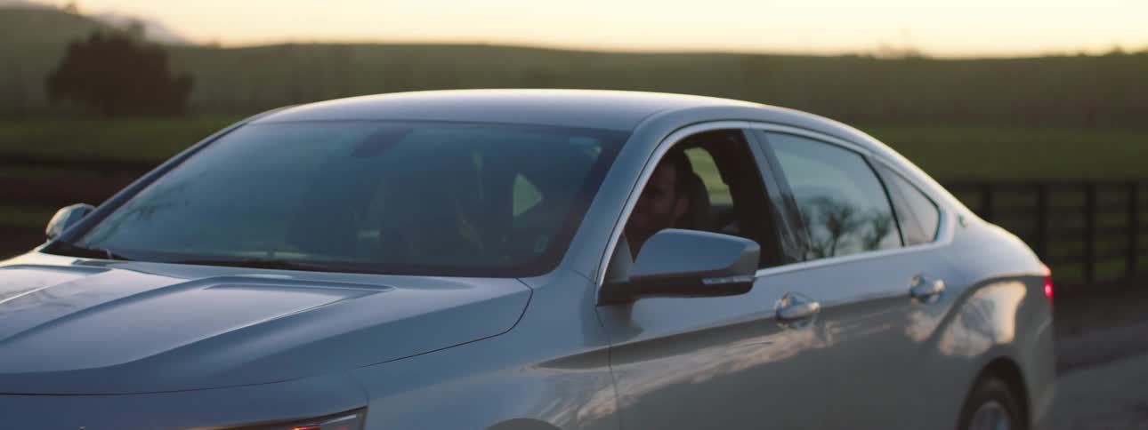After glow remains on horizon as man drives car past fields backed with forest and mountains