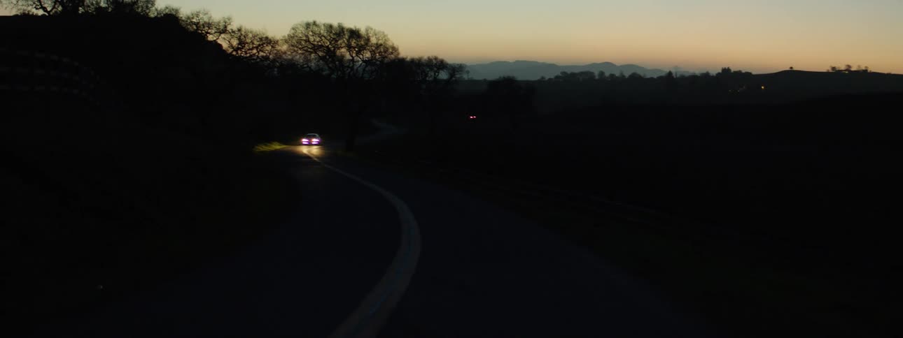 Car having its headlights on drives on a country road at dusk
