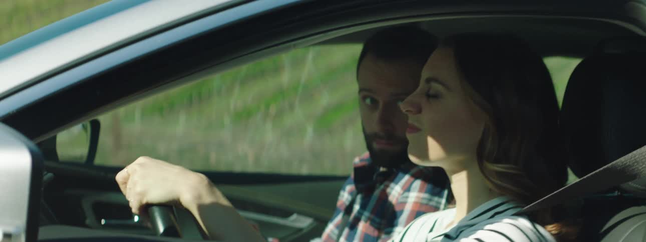 A woman drives a car past a grassland with a man in the passenger seat