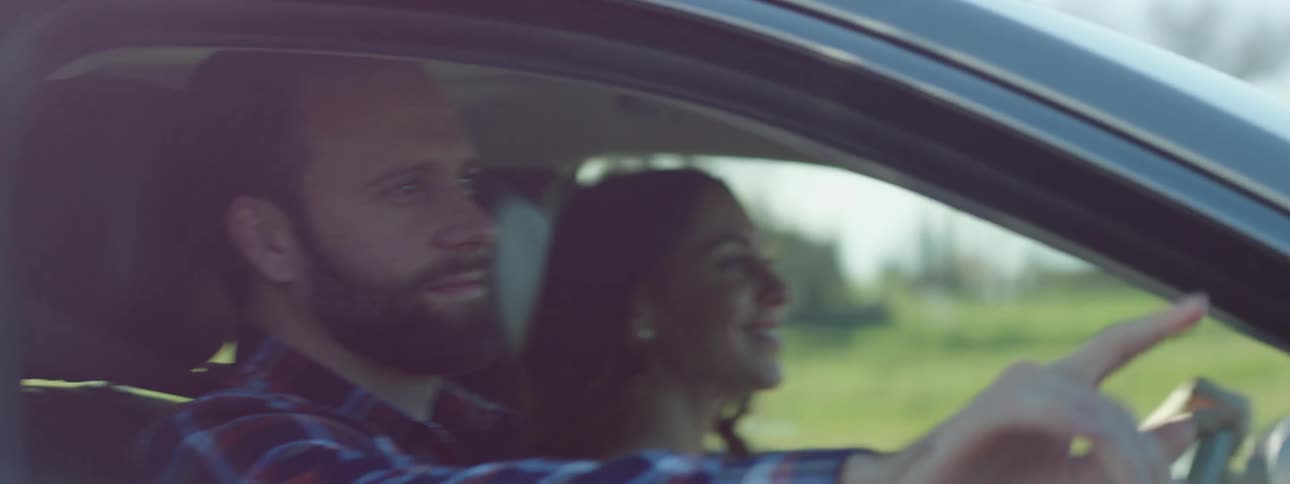A man and a woman driving in a car with grass trees and the sky in the background