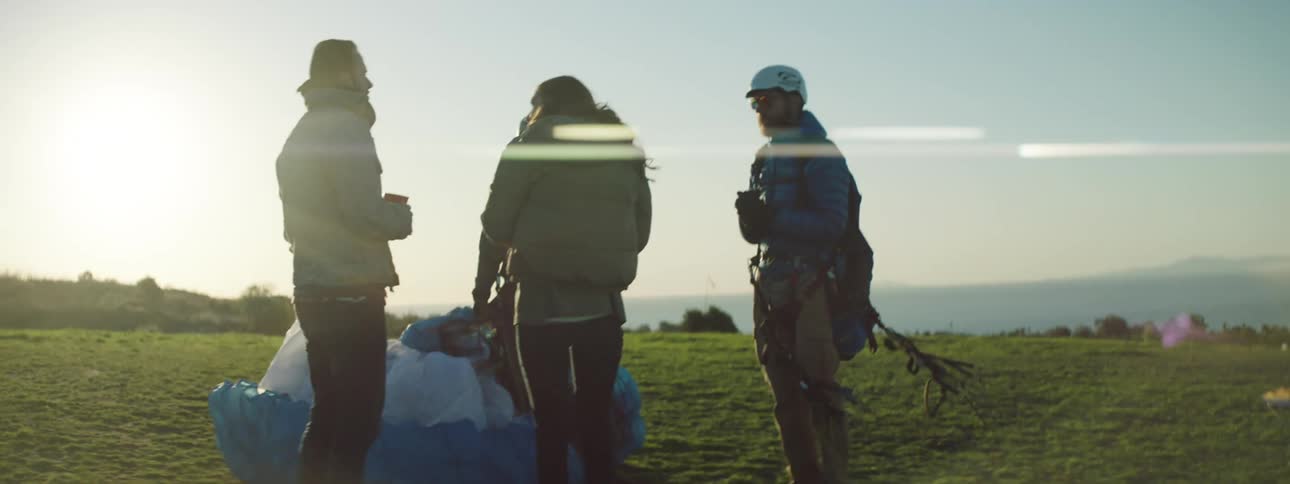 A pair of men in paragliding equipment speak to a man and woman holding cups as the four of them stand in a field