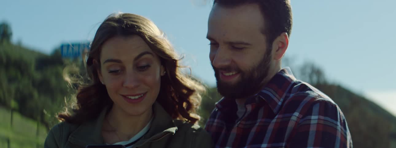 Man with a beard and striped shirt and woman with brown curly hair use a smartphone together in the countryside