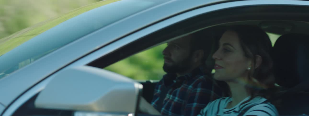 Woman with brown hair talks to man with a beard and a striped shirt while driving a grey car