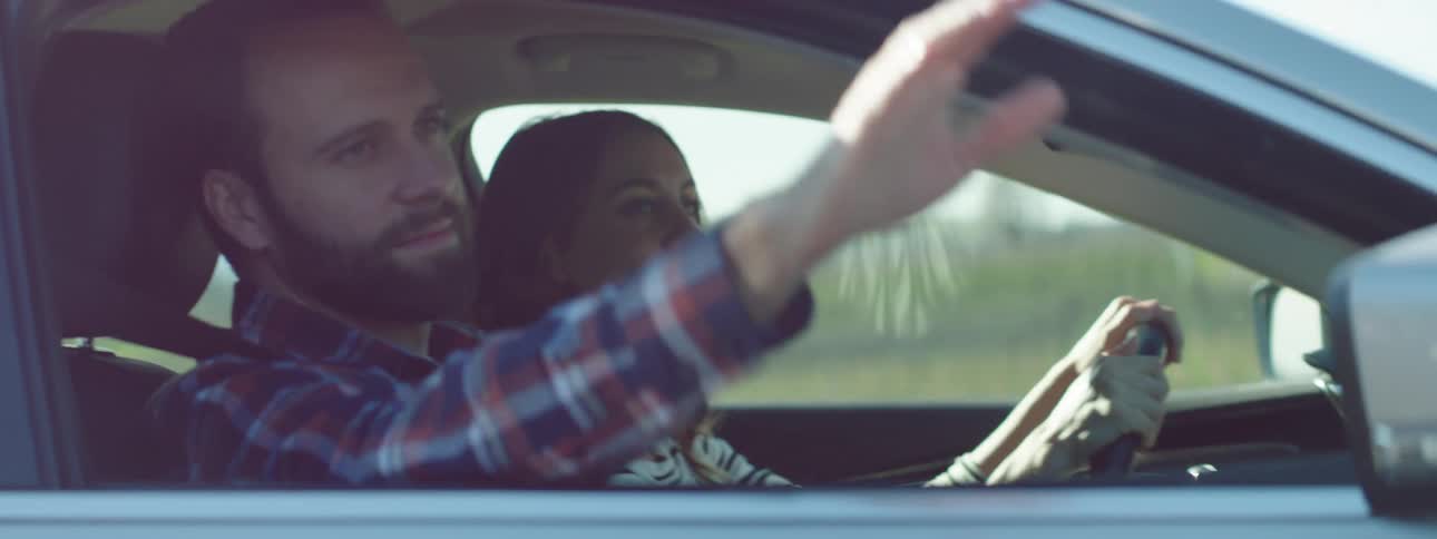 Man with a beard and a striped shirt sticks his hand out of the car window while woman drives the car