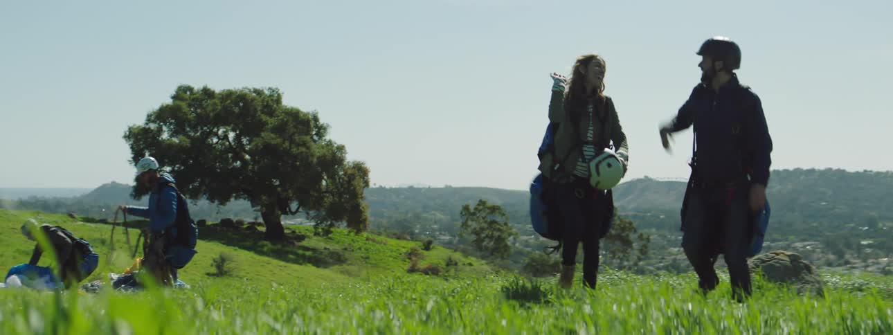 A man and a woman walk and high five outside on a grassy hill on a sunny day while two more people pick up parachutes behind them