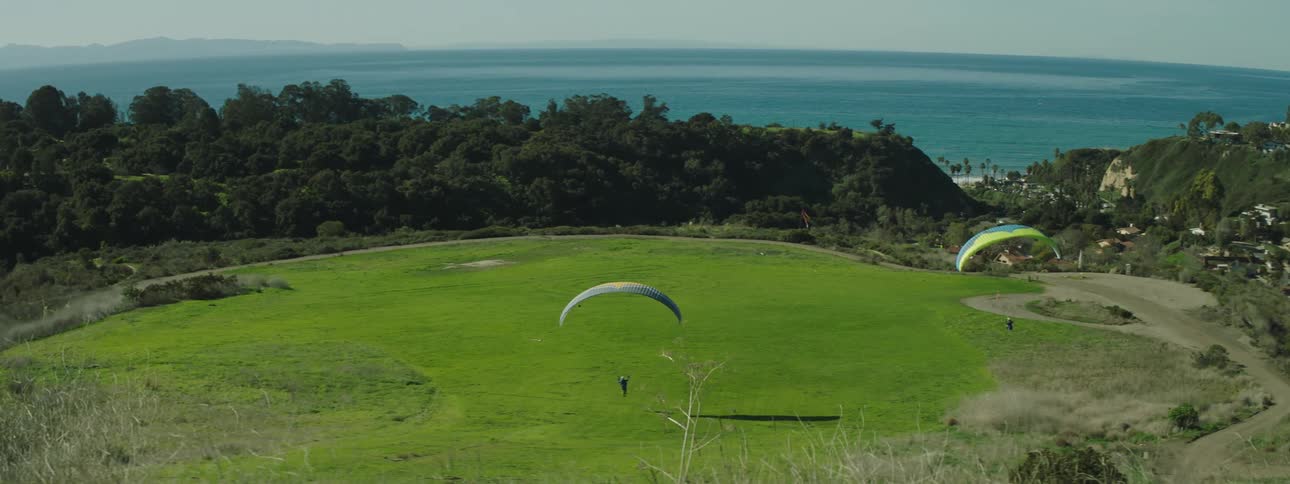 Two people parachute into an open grass field area ocean waters