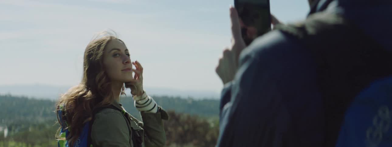 Woman stands in front of scenic view while man photographs her with mobile phone