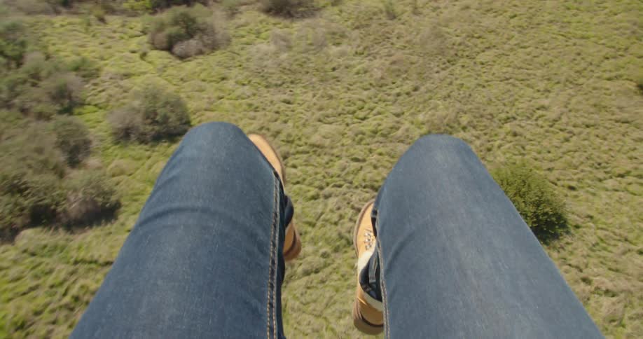 A persons legs with jeans and boots dangling in the air with grass and shrubs below