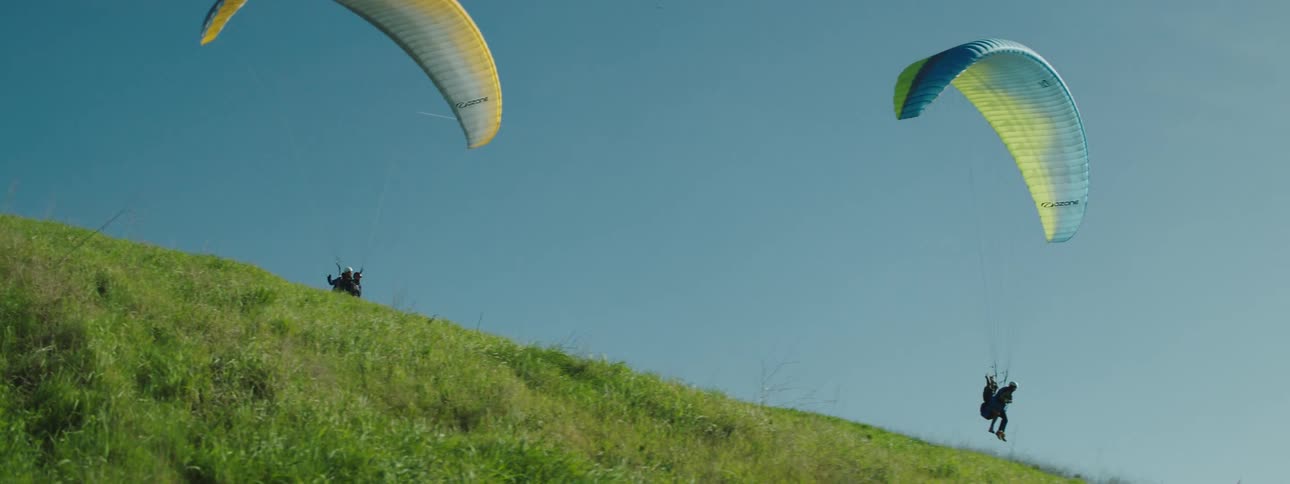 People ride paragliders in pairs over grass covered hill slope under a clear sky