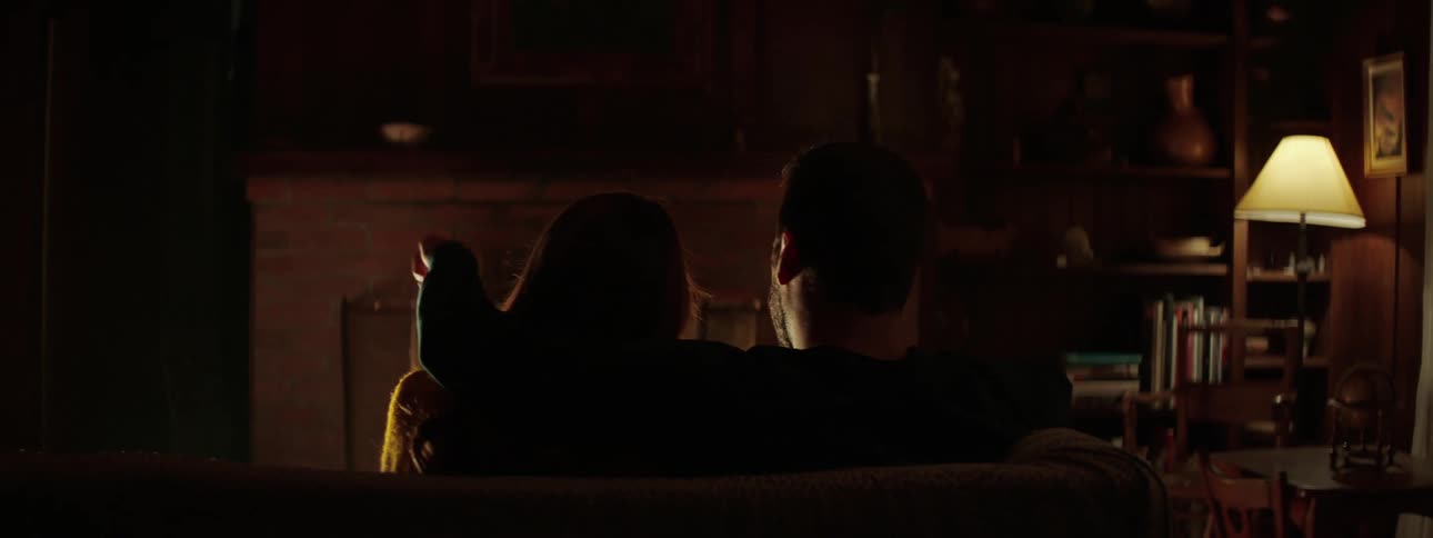 Male places his arm around females shoulder as they sit on one end of the sofa together and watch a roaring fire in the fireplace in a dimly lit family room