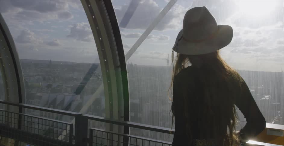 A woman is walking along a railing looking out a window over a city