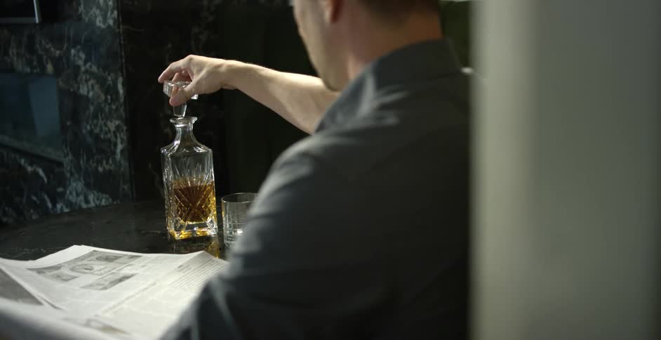 Man sitting at table reading newspaper prepares to pour alcohol into glass from carafe