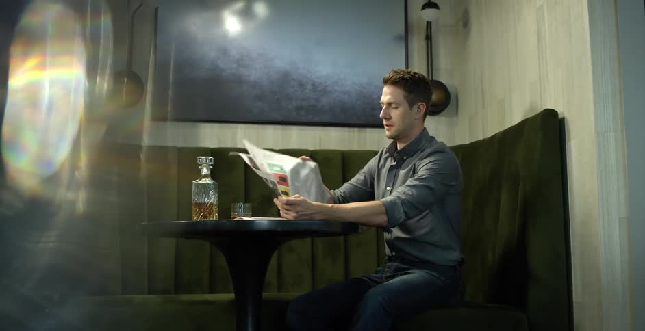 Man with a grey shirt and jeans sits on the couch and reads a newspaper in a cafe