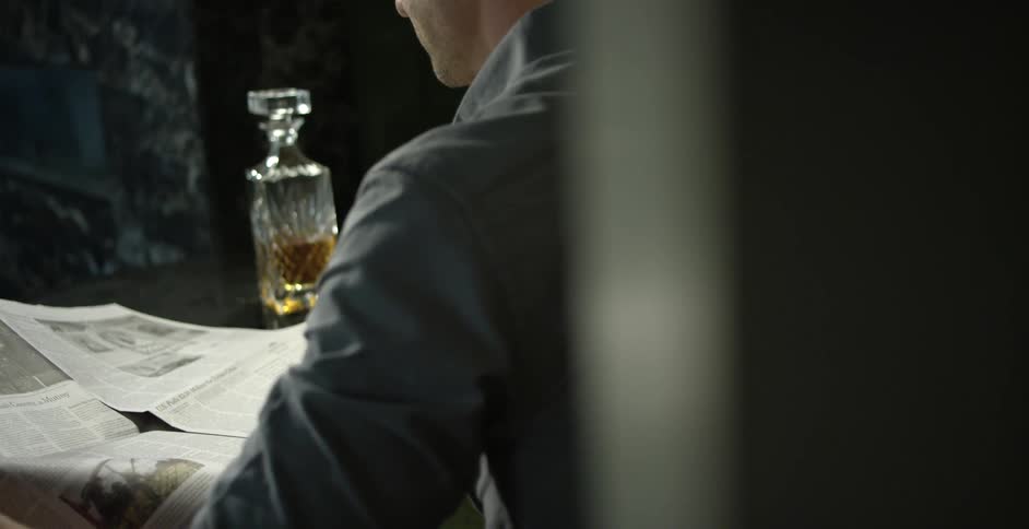 A man takes a drink of an alcoholic beverage before returning to reading a newspaper