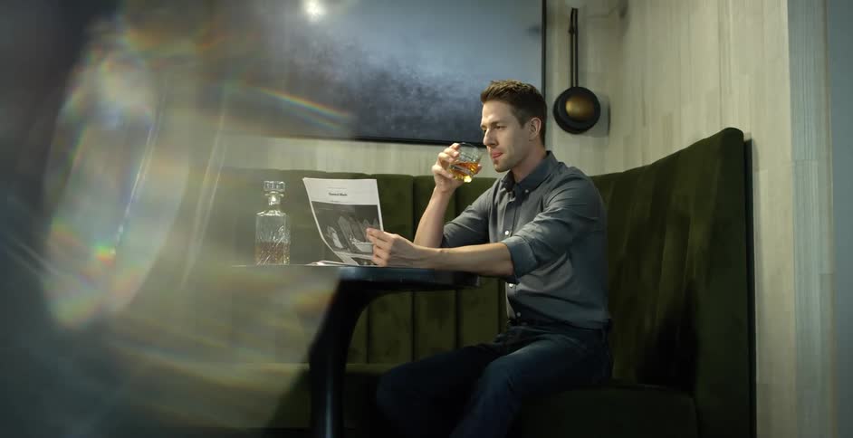 A man sits at a corner booth in a pub drinking a glass of liquor and reading a newspaper