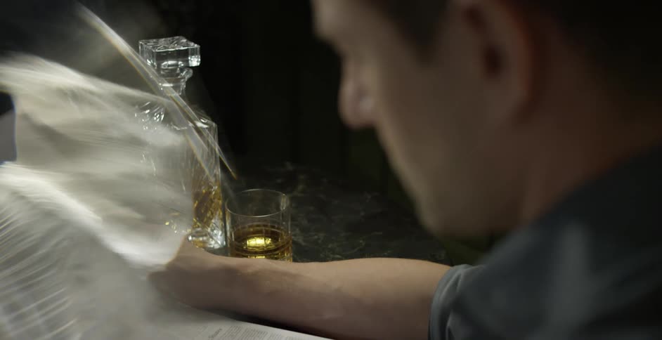 A man reads a newspaper while sitting at a table where a bottle of liquor and drinking glass are placed