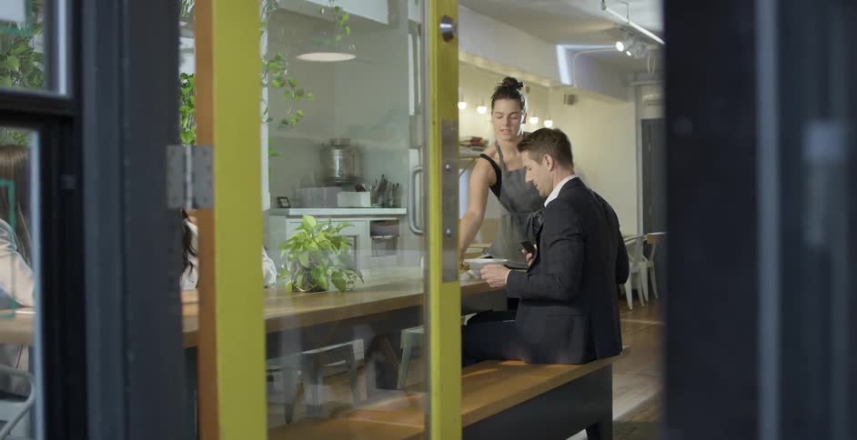 A female server places a bowl down in front of a man sitting at a bench in a small cafe
