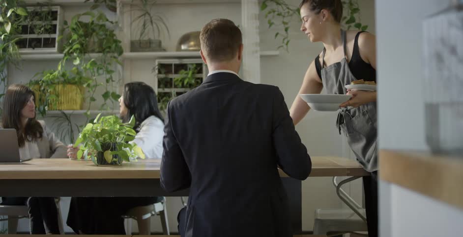 Man wearing a black suit sits at a table and waitress brings food and drink for the customer in a cafeteria