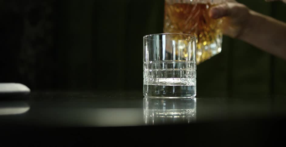 Man lifts a crystal bottle filled with whiskey and serves it on a glass over a table