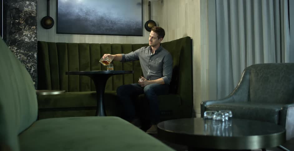 Man pours alcohol from a decanter into a glass as he sits on a corner sofa