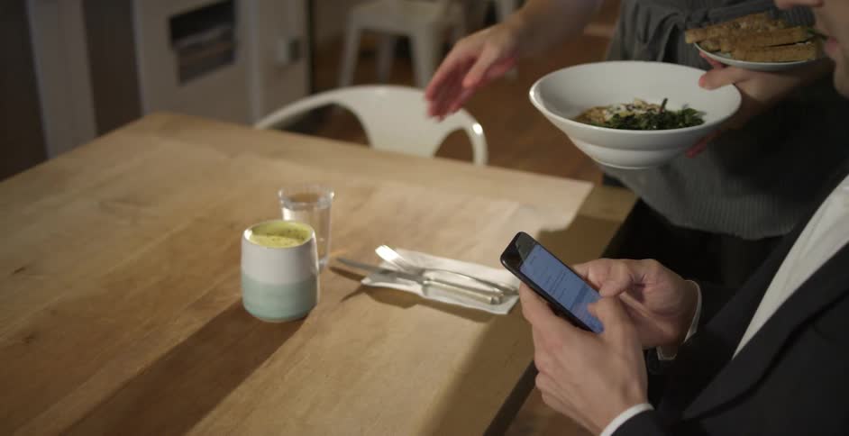 Businessman wearing a black suit sits at the table and uses a smartphone while waitress with a grey apron brings food and drink to the man at his table in a cafeteria