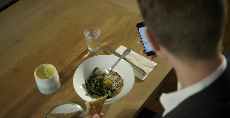 Man with a black suit sitting at a table eats food and uses a smartphone in a restaurant