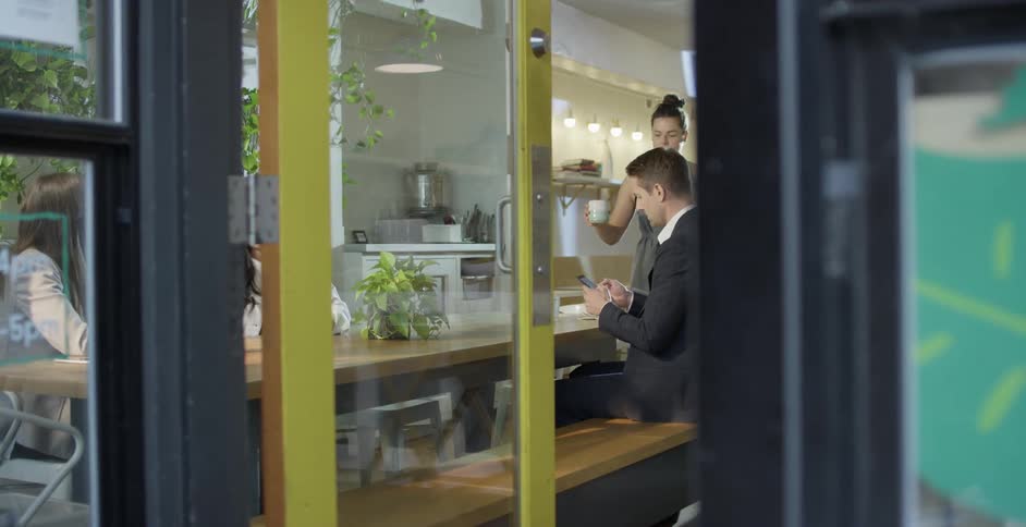 Man in black suit sits at a table and uses a smartphone as waitress with a grey apron brings drink and food to the customer at his table in a cafeteria